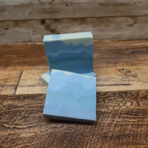 A handcrafted bar of soap with layered shades of blue and white, resembling a frosty winter landscape. The soap is displayed on a rustic wooden surface, highlighting its smooth texture and artisan design.