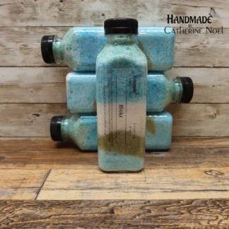 Bloke soaking salts in a clear plastic bottle with layers of earthy green and blue tones, labeled "Handmade by Catherine Noel," displayed on a rustic wooden surface.