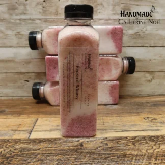 Frosted Whimsy soaking salts in a clear plastic bottle with delicate pink tones, labeled "Handmade by Catherine Noel," displayed on a rustic wooden surface.