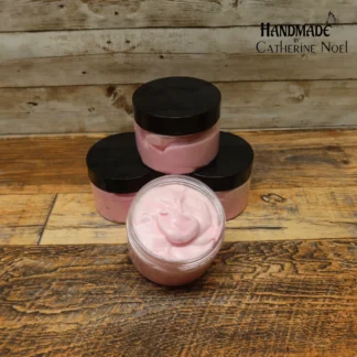 Frosted Whimsy whipped body butter with soft pink and white swirls, displayed in a clear jar with a black lid on a rustic wooden surface.