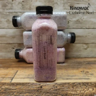 Love Bomb soaking salts in a clear plastic bottle with romantic lavender and pink tones, labeled "Handmade by Catherine Noel," displayed on a rustic wooden surface.