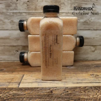 Peach Bliss soaking salts in a clear plastic bottle with soft peach tones, labeled "Handmade by Catherine Noel," displayed on a rustic wooden surface.