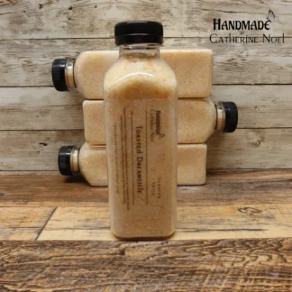 A clear plastic bottle filled with orange-toned Toasted Dreamsicle soaking salts, labeled with "Handmade by Catherine Noel," displayed in front of a rustic wooden background.