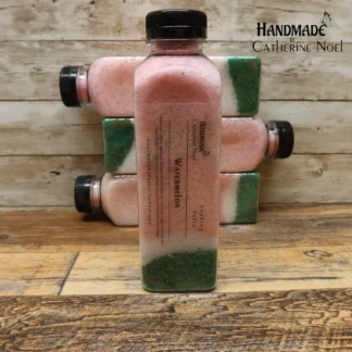 Watermelon soaking salts in a clear plastic bottle with pink and green layers, labeled "Handmade by Catherine Noel," displayed on a rustic wooden surface.