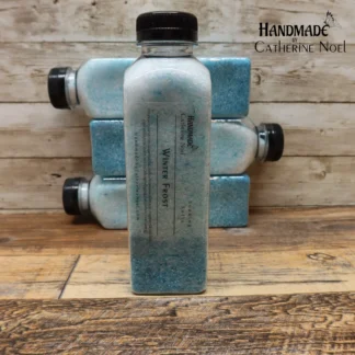 Winter Frost soaking salts in a clear plastic bottle with icy blue tones, labeled "Handmade by Catherine Noel," displayed on a rustic wooden surface.